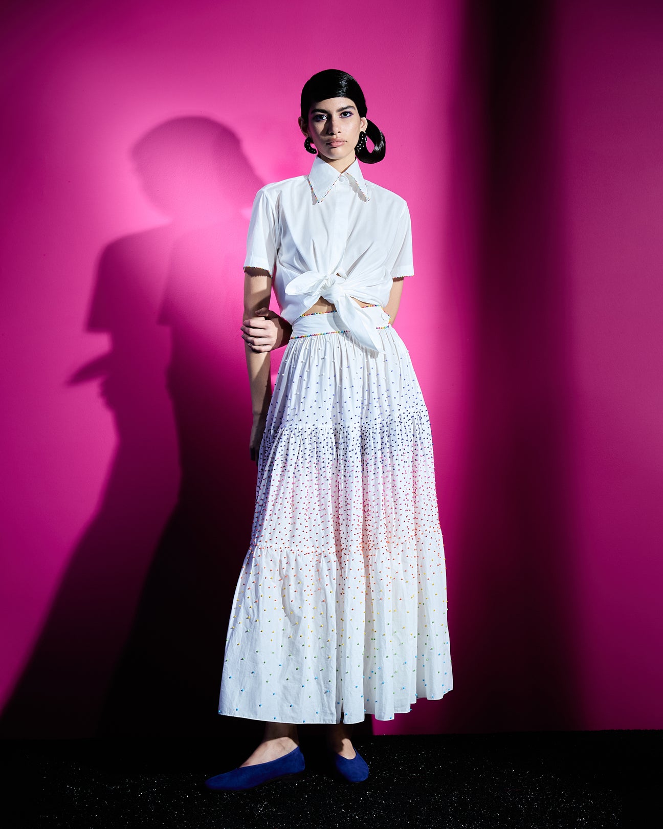 Modelo vestindo a Saia 'Leonora' Dots, em branco, é feita de algodão e possui cintura alta. Com um delicado bordado manual de miçangas coloridas, é ideal para composições elegantes e descontraídas. Combine a saia com a Camisa 'Rio' Dots para um look total branco com um toque de cor das miçangas; a camisa pode ser amarrada para um toque moderno e versátil.