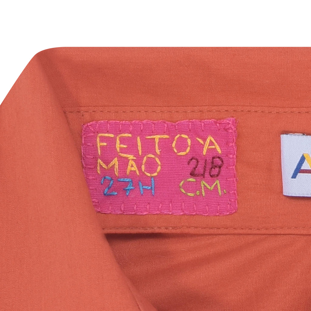 Etiqueta da Camisa Vera Listras laranja com inscrição “Feito à Mão” sobre tecido em tricoline de algodão.