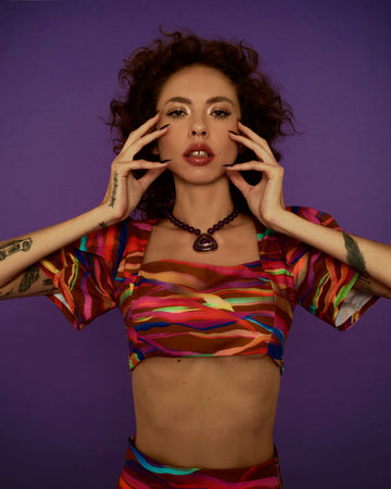 Modelo vestindo o Top 'Yasmin' Tigre da Alix. Top cropped com estampa listrada colorida sobre fundo marrom. Estilo feminino moderno.