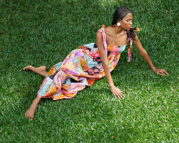 Modelo vestindo o Vestido 'Maria' Mar Rosa da Alix deitada no gramado. Peça longa com estampa multicolorida e alças com laço.